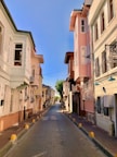 A narrow, paved street lined with pastel-colored buildings featuring bay windows, balconies, and iron window grilles. The street is empty, and the sky above is clear blue. Signs indicate potential guesthouses or businesses along the street.