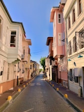 A narrow, paved street lined with pastel-colored buildings featuring bay windows, balconies, and iron window grilles. The street is empty, and the sky above is clear blue. Signs indicate potential guesthouses or businesses along the street.