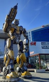 A large, yellow and black robot statue stands prominently in an urban area in front of a modern shopping center. The robot has a humanoid shape with intricate mechanical details and is posed holding an arm upwards. Surrounding it are retail stores like H&M and a shopping area bustling with activity.