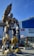 A large, yellow and black robot statue stands prominently in an urban area in front of a modern shopping center. The robot has a humanoid shape with intricate mechanical details and is posed holding an arm upwards. Surrounding it are retail stores like H&M and a shopping area bustling with activity.