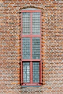 A tall, narrow window with a red frame set into a brick wall. The window is composed of multiple small square panes of glass, arranged in a grid pattern, typical of old or historic buildings.