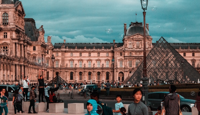 A bustling scene with people gathered around the glass pyramid entrance at the Louvre Museum. The historic architecture of the museum building is visible in the background under an overcast sky. The crowd includes tourists taking photos and sightseeing.
