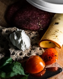A wooden surface displaying a selection of food items, including a slice of salami, a rolled piece of cheese with herbs, round crispbread topped with a dollop of creamy white cheese, and a halved tomato sprinkled with herbs. Basil leaves are also visible, adding a touch of green.