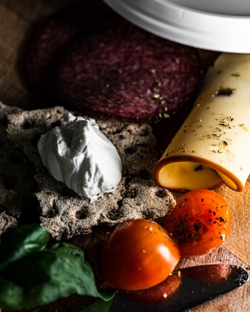 A wooden surface displaying a selection of food items, including a slice of salami, a rolled piece of cheese with herbs, round crispbread topped with a dollop of creamy white cheese, and a halved tomato sprinkled with herbs. Basil leaves are also visible, adding a touch of green.