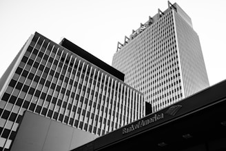 Black and white photograph of tall, modern buildings with vertical and horizontal lines. The architecture features a prominent grid pattern, and there's a visible logo on the tallest building.