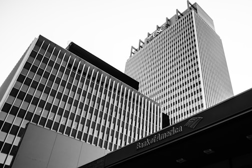 Black and white photograph of tall, modern buildings with vertical and horizontal lines. The architecture features a prominent grid pattern, and there's a visible logo on the tallest building.