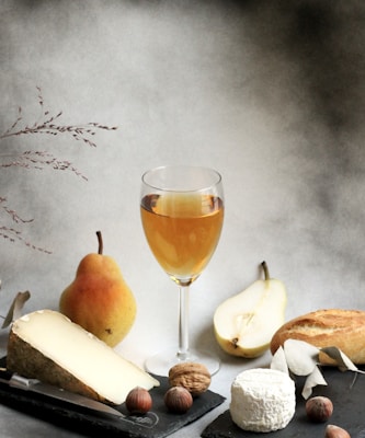 A still life arrangement featuring a glass of amber-colored wine surrounded by various foods. Visible items include a whole pear, a pear half, a large wedge of cheese with a knife, a round soft cheese, a walnut, hazelnuts, and a piece of baguette. The background is an artistic, softly blurred backdrop that adds a classic and timeless feel.
