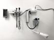 Medical equipment is neatly mounted on a white wall, including an otoscope, an ophthalmoscope, a sphygmomanometer, and a thermometer. The layout suggests a clinical setting likely intended for patient examination.