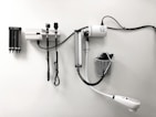 Medical equipment is neatly mounted on a white wall, including an otoscope, an ophthalmoscope, a sphygmomanometer, and a thermometer. The layout suggests a clinical setting likely intended for patient examination.