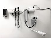 Medical equipment is neatly mounted on a white wall, including an otoscope, an ophthalmoscope, a sphygmomanometer, and a thermometer. The layout suggests a clinical setting likely intended for patient examination.