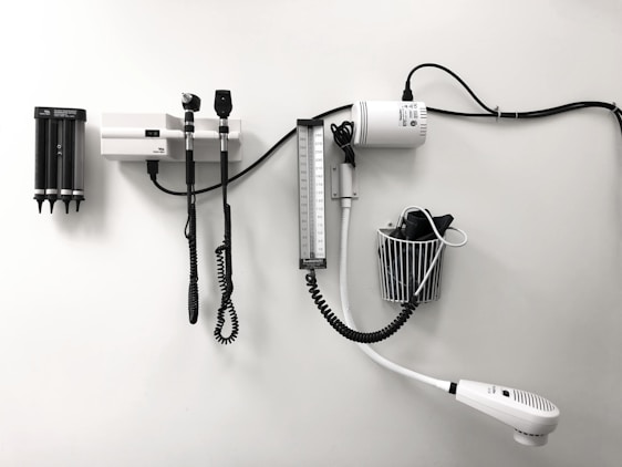 Medical equipment is neatly mounted on a white wall, including an otoscope, an ophthalmoscope, a sphygmomanometer, and a thermometer. The layout suggests a clinical setting likely intended for patient examination.