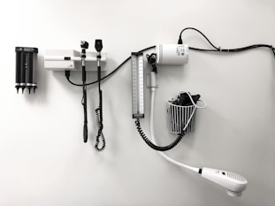Medical equipment is neatly mounted on a white wall, including an otoscope, an ophthalmoscope, a sphygmomanometer, and a thermometer. The layout suggests a clinical setting likely intended for patient examination.