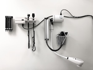 Medical equipment is neatly mounted on a white wall, including an otoscope, an ophthalmoscope, a sphygmomanometer, and a thermometer. The layout suggests a clinical setting likely intended for patient examination.