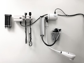 Medical equipment is neatly mounted on a white wall, including an otoscope, an ophthalmoscope, a sphygmomanometer, and a thermometer. The layout suggests a clinical setting likely intended for patient examination.