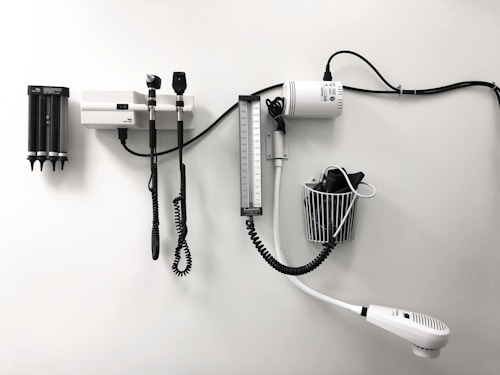 Medical equipment is neatly mounted on a white wall, including an otoscope, an ophthalmoscope, a sphygmomanometer, and a thermometer. The layout suggests a clinical setting likely intended for patient examination.