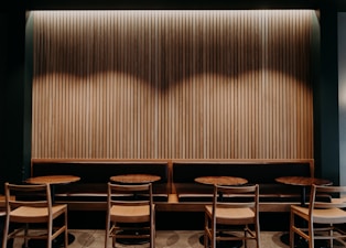 A cozy, dimly lit seating area with wooden chairs and round tables arranged in front of a long bench. The background features a vertically-paneled wooden wall with a strip of light illuminating the upper section, creating a warm and inviting atmosphere.