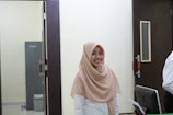 A woman wearing a light brown hijab and a white shirt is smiling while standing in an office-like environment. She is situated between a dark door and a computer monitor, with a grey cabinet and a trash bin visible in the background.
