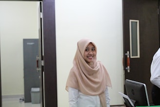 A woman wearing a light brown hijab and a white shirt is smiling while standing in an office-like environment. She is situated between a dark door and a computer monitor, with a grey cabinet and a trash bin visible in the background.