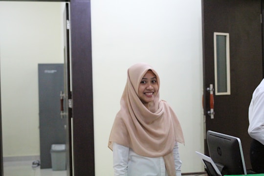 A woman wearing a light brown hijab and a white shirt is smiling while standing in an office-like environment. She is situated between a dark door and a computer monitor, with a grey cabinet and a trash bin visible in the background.