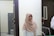 A woman wearing a light brown hijab and a white shirt is smiling while standing in an office-like environment. She is situated between a dark door and a computer monitor, with a grey cabinet and a trash bin visible in the background.