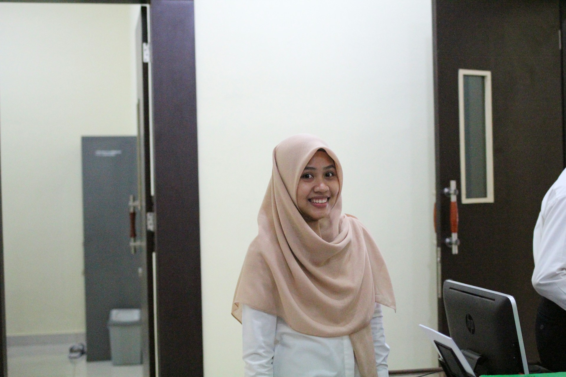 A woman wearing a light brown hijab and a white shirt is smiling while standing in an office-like environment. She is situated between a dark door and a computer monitor, with a grey cabinet and a trash bin visible in the background.