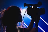 A silhouetted figure, possibly a child, is operating a video camera with a bright, colorful background illuminated by blurred lighting lines.