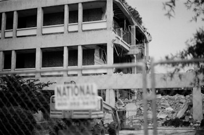 A partially demolished building with visible debris and rubble. The structure appears to be mid-demolition, with exposed floors and wires hanging. In the foreground, a chain-link fence is present, and a sign marked 'NATIONAL' is attached to it.
