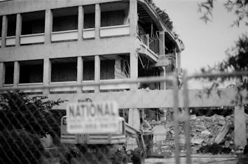 A partially demolished building with visible debris and rubble. The structure appears to be mid-demolition, with exposed floors and wires hanging. In the foreground, a chain-link fence is present, and a sign marked 'NATIONAL' is attached to it.