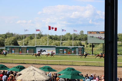 A horse racing event is taking place on a sunny day, with a racecourse surrounded by greenery. Several horses with jockeys are actively racing across the track. Spectators are gathered under shaded canopies in the foreground. Multiple flags, including a prominent Canadian flag, are flying above a scoreboard, and a large screen displays the race.