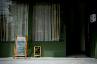 A street-level window with closed beige curtains is flanked by two signs on the sidewalk. The larger sign is wooden with glass, displaying information about coffee being served upstairs from morning to evening. The smaller chalkboard sign advertises craft beer, wine, and cocktails available on Tuesdays and Fridays. Both signs are angled for visibility, against a green wall backdrop.