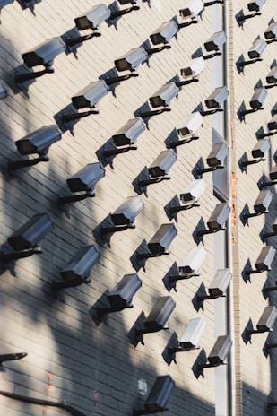 A wall covered in numerous surveillance cameras, all aligned in neat rows and columns. The arrangement creates a pattern against the light-colored brick wall, casting strong shadows that add a sense of repetition and uniformity.