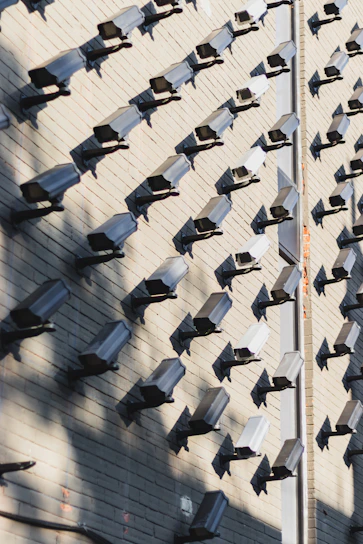 A wall covered in numerous surveillance cameras, all aligned in neat rows and columns. The arrangement creates a pattern against the light-colored brick wall, casting strong shadows that add a sense of repetition and uniformity.