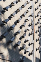 A wall covered in numerous surveillance cameras, all aligned in neat rows and columns. The arrangement creates a pattern against the light-colored brick wall, casting strong shadows that add a sense of repetition and uniformity.