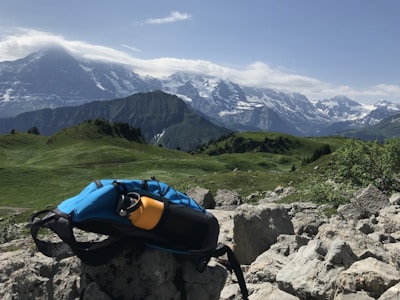 A vibrant landscape featuring rugged mountains partially covered in snow under a clear blue sky. Lush green hills roll across the foreground, dotted with rocks and sparse vegetation. A backpack and a yellow water bottle are visible, resting on a rock, suggesting a hiking or outdoor activity setting.