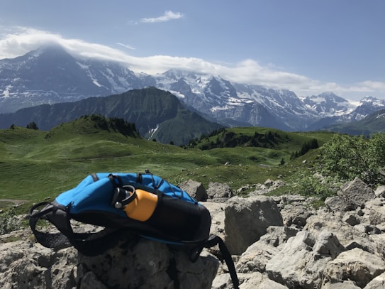 A vibrant landscape featuring rugged mountains partially covered in snow under a clear blue sky. Lush green hills roll across the foreground, dotted with rocks and sparse vegetation. A backpack and a yellow water bottle are visible, resting on a rock, suggesting a hiking or outdoor activity setting.