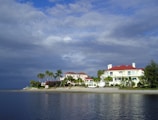 A large, luxurious house with red roofs sits along a tranquil waterfront, surrounded by palm trees and lush greenery. The sky is partly cloudy, casting a mix of shadows and light on the scene.