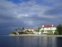 A large, luxurious house with red roofs sits along a tranquil waterfront, surrounded by palm trees and lush greenery. The sky is partly cloudy, casting a mix of shadows and light on the scene.