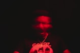 A blurry image displays a person with a distorted face, enveloped in a red hue, creating a sense of motion or vibration. The person wears a dark shirt with a graphic design featuring several faces and has a chain necklace.