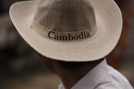 A person wearing a straw hat with the word 'Cambodia' written on the band. The person is seen from the back and is wearing a white shirt. The background is blurred and indistinct.