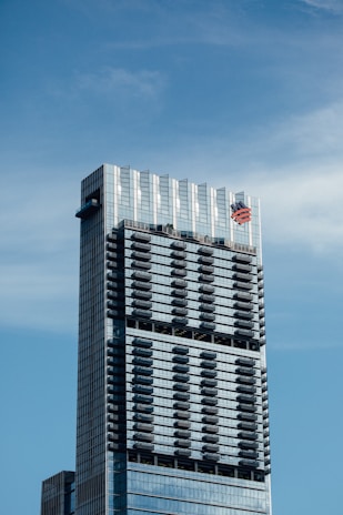 A tall, modern skyscraper with a sleek glass exterior, featuring multiple balconies evenly distributed across its facade. The building has a prominent logo near the top.