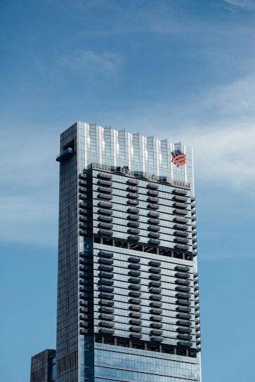 A tall, modern skyscraper with a sleek glass exterior, featuring multiple balconies evenly distributed across its facade. The building has a prominent logo near the top.