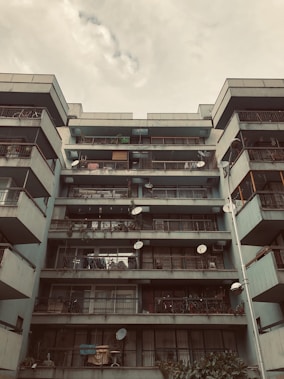 An urban apartment building with multiple balconies, each adorned with various items like satellite dishes, plants, and clotheslines. The structure has a modernist architectural style, and the sky above is cloudy.