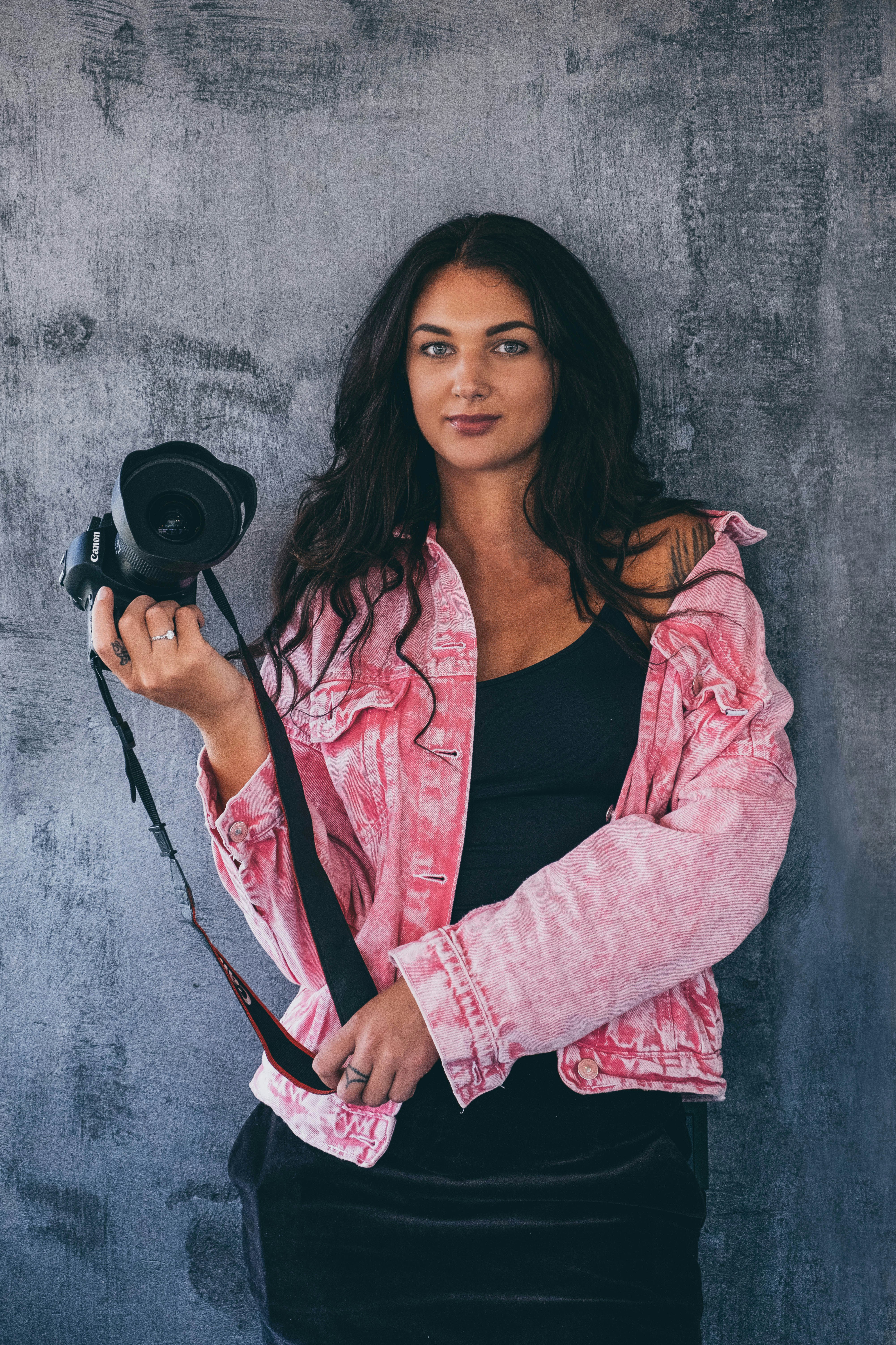 woman standing in front of gray wall holding black DSLR camera