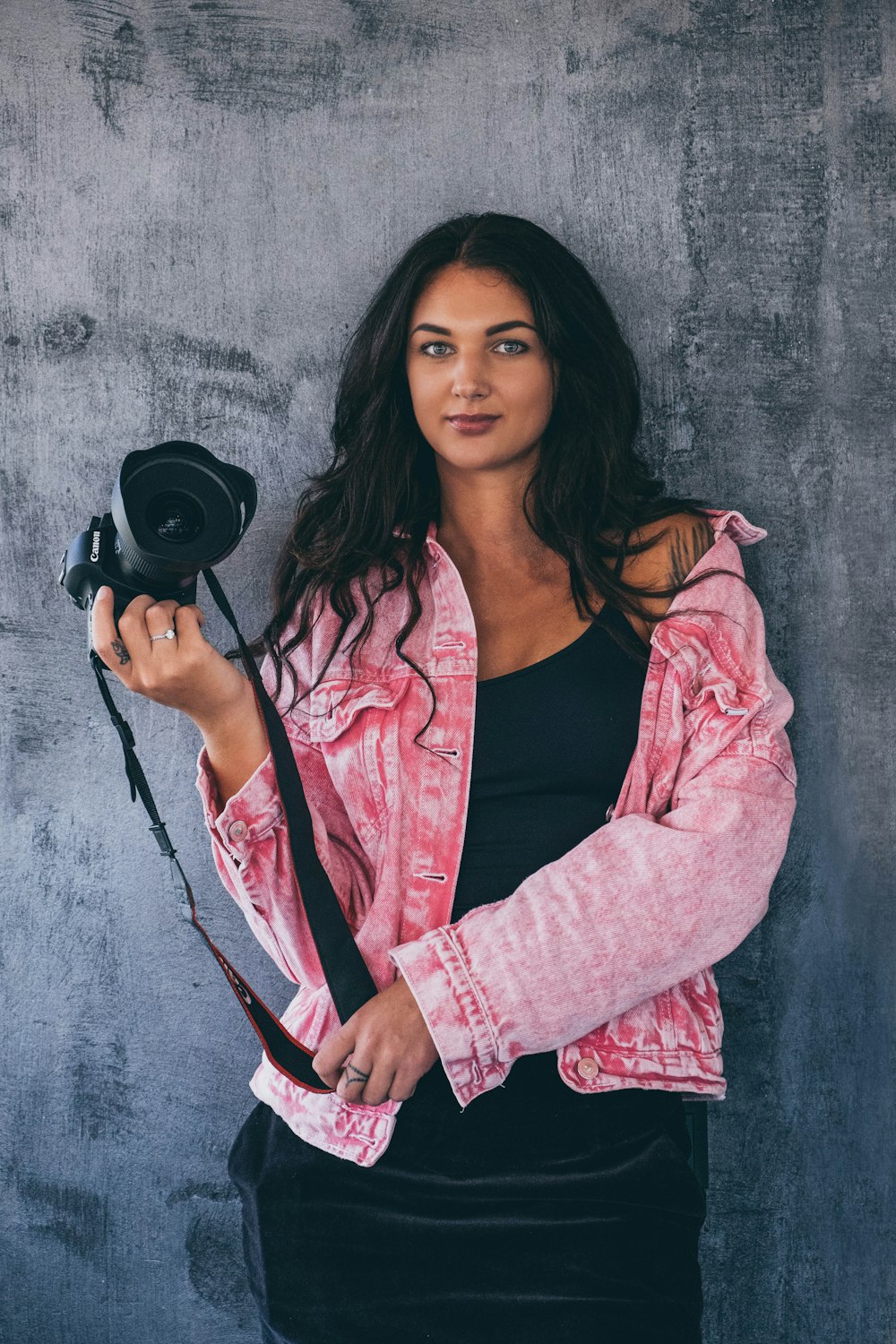 woman standing in front of gray wall holding black DSLR camera