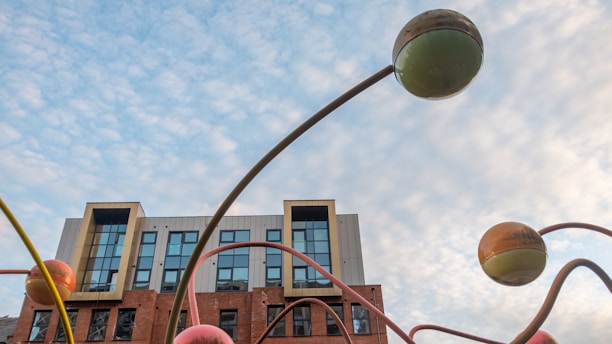 A building with a modern design featuring large windows and a combination of brick and concrete sections. In the foreground, there are large, colorful, spherical sculptures mounted on thin, curved poles, set against a partly cloudy sky.