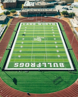 Aerial view of a football field with clearly marked yard lines and end zones labeled with 'LAKE WORTH' and 'BULLFROGS'. The field is surrounded by a red running track, and in the background, there are school buildings and parked vehicles.