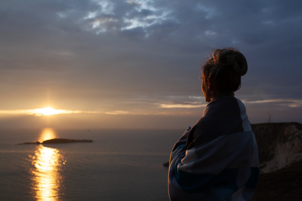 woman wrapped in blanket standing on higher ground watching sunset over the sea