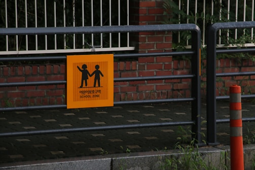 A metal railing with a bright orange sign hung on it, displaying silhouettes of a child and an adult holding hands accompanied by text in Korean and English indicating a school zone. Below, concrete blocks form a pathway adjacent to a brick wall partially covered by foliage. A traffic cone with orange and white stripes stands nearby.