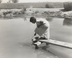 A person crouches on a wooden plank extending over a body of water, reaching down with a pan or container to collect water. The scene is set in a rural area with open fields in the background and large hay bales nearby.