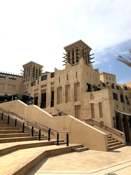 A large multi-level building with stairs leading up to a structure that features traditional Middle Eastern architectural elements. The design includes beige stone walls, wooden beams, and wind towers. A clear sky and a palm tree are visible in the background.